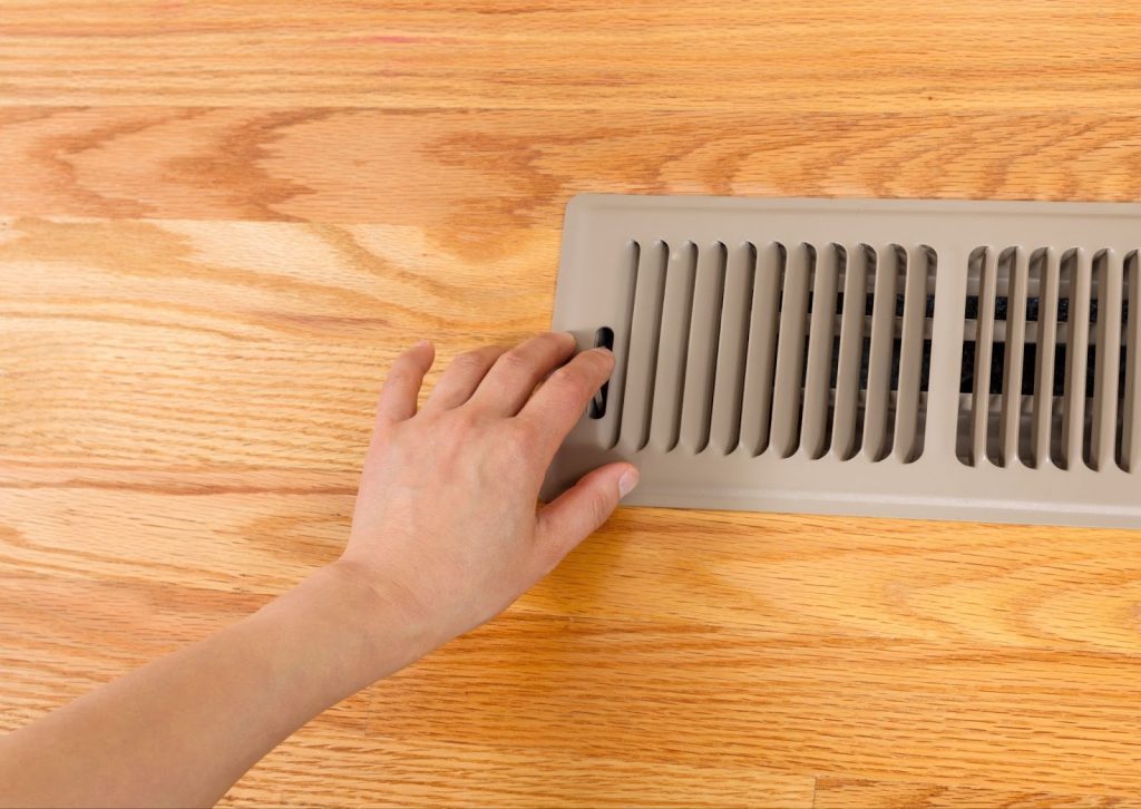 Person adjusting air vent on wooden floor, comparing radiant heating and traditional forced air systems