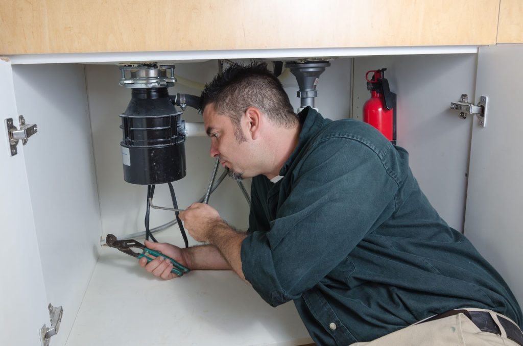 A professional plumber fixing a sink under a cabinet. He is working on garbage disposal repairs.