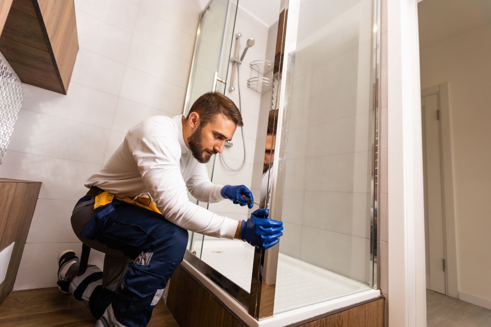 A plumber in a white shirt and blue gloves repairs a shower door, ensuring quality service for your bathtub or shower needs.