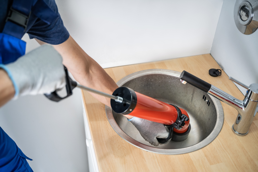 A man in blue overalls employs a red tool to clean a sink, highlighting the importance of regular household upkeep.